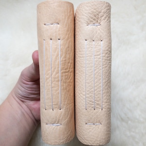 May include: Two beige leather-bound journals with visible stitching along the spine. The journals are held upright, showcasing their textured covers and the detail of the stitching. The color is a light, natural beige.
