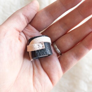 May include: A black leather ring with a white leather accent. The ring is wrapped around a finger.