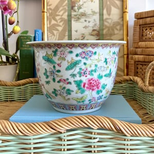 May include: A white ceramic planter with a flared rim, adorned with a colorful floral pattern featuring pink peonies, blue and yellow flowers, and green leaves. The planter sits on a blue surface within a woven basket.