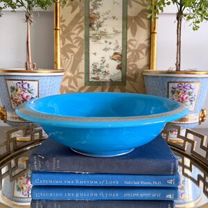 May include: A bright turquoise ceramic bowl with a gold-toned laurel leaf pattern along the rim, resting on a stack of three dark blue hardcover books titled Catching the Rhythm of Love by Neil Clark Warren, Ph.D. with gold lettering.