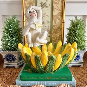 May include: A white ceramic monkey figurine holding a banana sits above a yellow and green banana-shaped bowl. The bowl rests on two books, with small green plants in blue and white pots on either side.