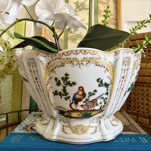 May include: A white porcelain bowl with a scalloped rim and gold trim, featuring a hand-painted scene of two birds perched on a branch amidst green foliage. The bowl has a flared base and decorative gold filigree patterns along the sides.