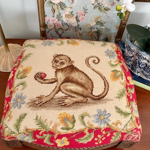 May include: A square needlepoint footstool cushion featuring a central brown monkey holding a small red fruit. The cream-colored center is surrounded by a decorative border of colorful flowers and green leaves on a vibrant red background.