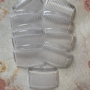 May include: A collection of clear plastic hair combs. Each comb has a curved top and a series of teeth. The combs are arranged on a white lace fabric with floral accents.