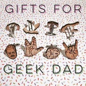 Peut inclure: Un ensemble de huit boutons de manchette en bois avec différents motifs sur le thème geek, notamment un salut vulcain de Star Trek, un dé de Donjons et Dragons, un geste de la main rock and roll et un portrait d'un homme avec des lunettes. Les boutons de manchette sont disposés sur un fond blanc avec des confettis colorés.