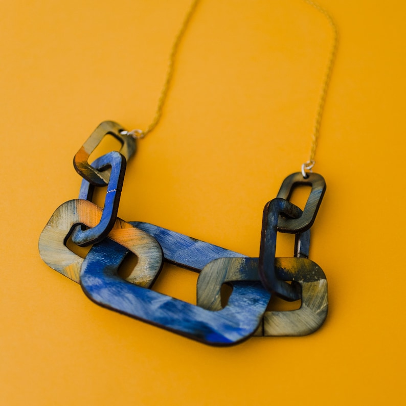 Op de afbeelding: Een chunky statement ketting met grote, in elkaar grijpende schakels die in tinten blauw en geel zijn geverfd. De ketting is gemaakt van hout en heeft een gouden ketting.