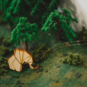 May include: A miniature wooden elephant figurine with a light brown finish, placed on a green moss-covered surface with miniature trees and a wooden fence in the background.
