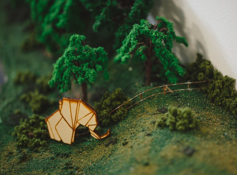 May include: A miniature wooden elephant figurine with a light brown finish, placed on a green moss-covered surface with miniature trees and a wooden fence in the background.