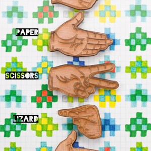 Peut inclure: Cinq découpes de mains en bois représentant les cinq gestes de la main du jeu Pierre Papier Ciseaux Lézard Spock. Les découpes de mains sont disposées verticalement de haut en bas avec les mots "PIERRE", "PAPIER", "CISEAUX", "LÉZARD" et "SPOCK" écrits sous chaque découpe. Les découpes sont sur un fond blanc avec un motif quadrillé de carrés verts, bleus et jaunes.