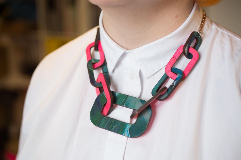Op de afbeelding: Een dikke statementketting gemaakt van grote, in elkaar grijpende schakels. De schakels zijn gemaakt van donkergroen en roze geverfd hout. De ketting is een gedurfd en opvallend accessoire.