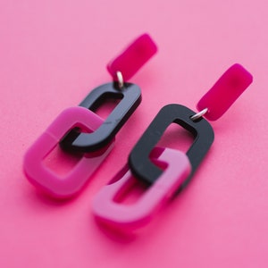 May include: A pair of black and pink acrylic earrings with a chain link design. The earrings are dangling and have a simple, modern style.