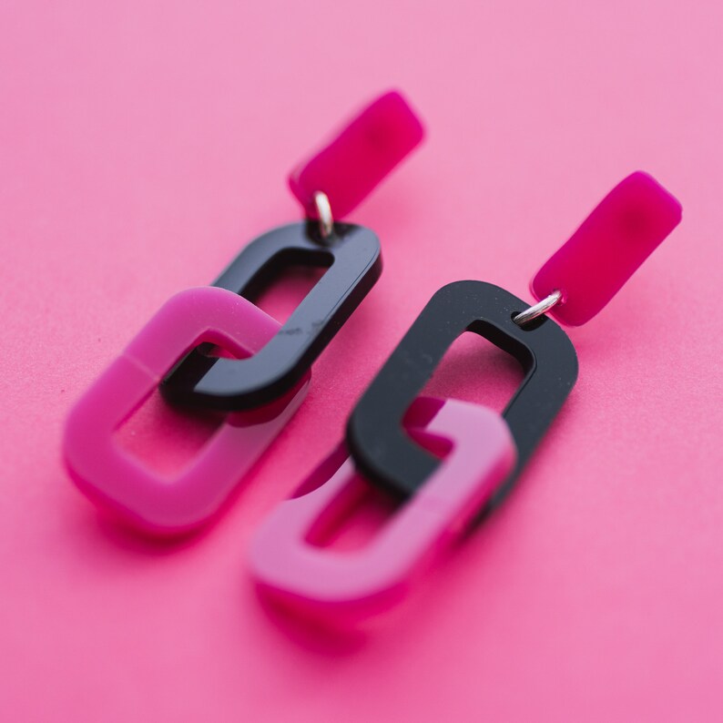 May include: A pair of black and pink acrylic earrings with a chain link design. The earrings are dangling and have a simple, modern style.