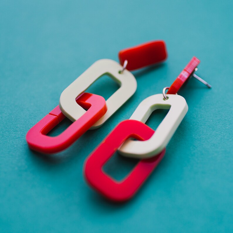 May include: A pair of red and cream colored plastic earrings shaped like linked chains. The earrings are on a blue background.