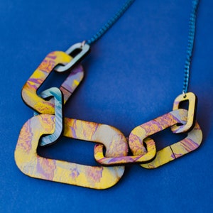 Op de afbeelding: Een kleurrijke ketting gemaakt van met elkaar verbonden houten stukken die in tinten geel, paars en blauw zijn geverfd. De ketting bestaat uit vijf schakels en ligt op een blauwe achtergrond.