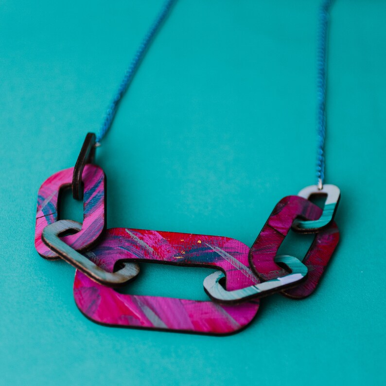 Op de afbeelding: Een roze en blauwe geschilderde houten ketting met een kettingschakelontwerp. De ketting ligt op een turquoise achtergrond.