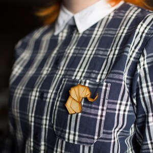 May include: A wooden elephant pin in a natural wood colour, worn on a blue and white checked shirt.