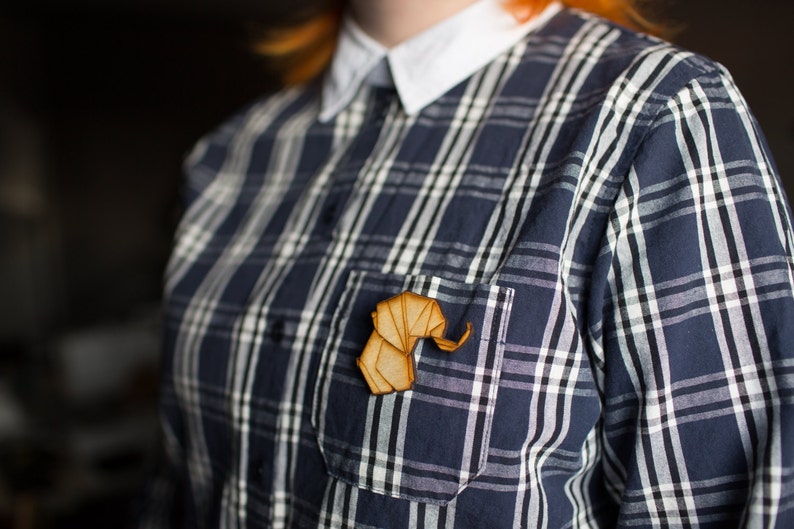 May include: A wooden elephant pin in a natural wood colour, worn on a blue and white checked shirt.