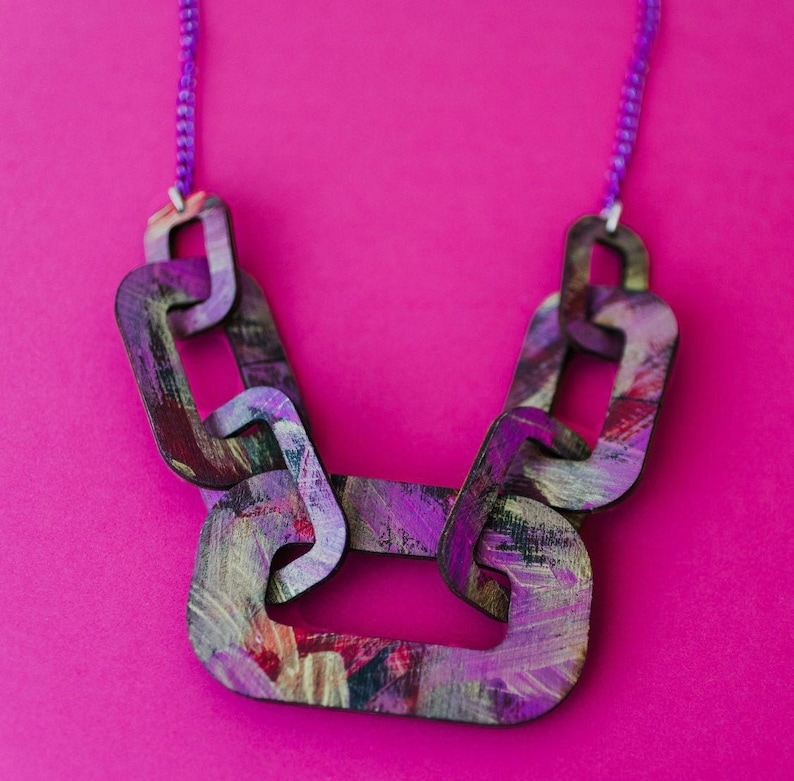 Op de afbeelding: Een dikke statementketting gemaakt van geschilderde houten schakels. De schakels zijn geschilderd in tinten paars, roze en goud en hebben een textuur. De ketting ligt op een roze achtergrond.