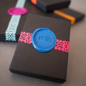 May include: A black gift box with a blue wax seal that says "Privy" in white lettering. The box is decorated with pink and blue paper lace.
