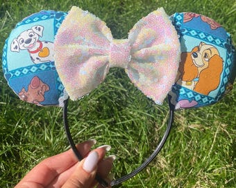 May include: A pair of black headband ears with a large sequined bow. The ears are decorated with images of cartoon dogs on a blue background. The bow is white with iridescent sequins.