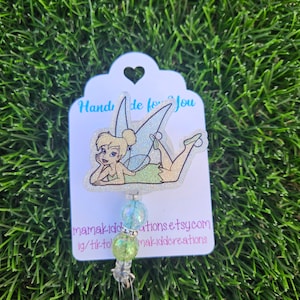 May include: A white card with a heart-shaped cutout at the top. The card has the text "Hand made for you" printed on it. A glitter-covered image of Tinkerbell is on the card. A green and blue beaded retractable badge holder is attached to the card. The text "mamakiddo creations etsy.com ig/tiktok mamakiddocreations" is printed on the card.