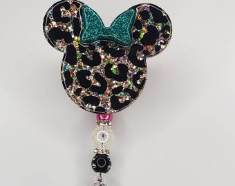 May include: A black and gold glitter leopard print Minnie Mouse head with a teal bow, attached to a retractable badge holder with pink, black, and white beads.