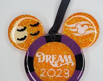 May include: Orange and purple glitter Mickey Mouse ornament with a black ribbon. The ornament has a purple circle with a black border and the words "DREAM 2023" in white. One ear has a white moon and bats, the other ear has a white Mickey Mouse head with a ship's wheel.