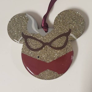 Peut inclure: Ornement Mickey Mouse en paillettes dor&eacute;es avec un motif rouge et marron de lunettes et d'un n&oelig;ud. L'ornement est dot&eacute; d'un ruban bordeaux.