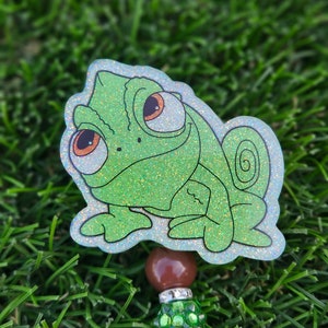 May include: A green glitter chameleon sticker with large eyes and a curled tail. The sticker is attached to a brown bead and a green beaded charm.
