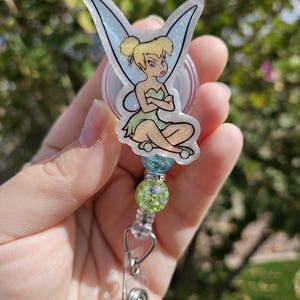 May include: A clear badge reel with a green and blue glitter charm featuring Tinkerbell from Disney's Peter Pan. The charm is a printed image of Tinkerbell sitting with her arms crossed, with a white glitter background.