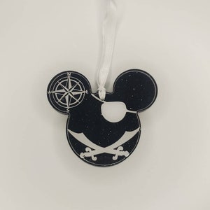 May include: Black Mickey Mouse-shaped ornament with a pirate theme. The ornament features a compass rose on one ear, an eye patch, and crossed swords. A white ribbon is attached for hanging.