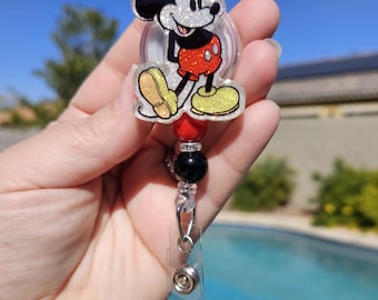 May include: A clear retractable badge holder with a glitter-covered Mickey Mouse character charm. The charm features a red and black Mickey Mouse design with yellow shoes. The holder has a black bead and a red bead with rhinestones.