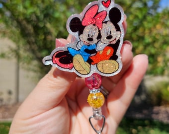 May include: A clear glitter charm with a cartoon image of Mickey Mouse and Minnie Mouse hugging. The charm is attached to a pink and yellow beaded retractable badge holder.