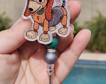 May include: A sparkly silver and green retractable badge holder with a glitter-covered Slinky Dog charm. The charm features a cartoon image of Slinky Dog from the Toy Story movies.