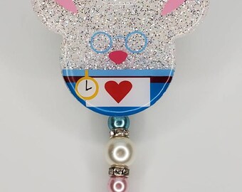 May include: White glitter resin badge reel with a white rabbit design. The rabbit has pink ears, blue glasses, a blue vest, a pocket watch, and a red heart. The reel has a white pearl, a pink pearl, and a blue rhinestone.