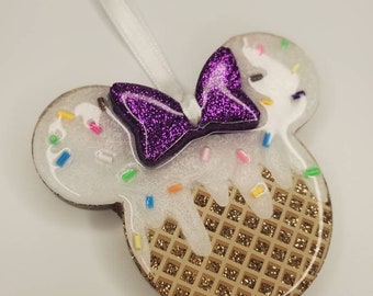 Könnte beinhalten: Ein weißer und goldener Glitzer-Harzornament in Form von Mickeys Kopf mit einer Schleife aus violettem Glitzer. Das Ornament hat eine Waffelkegeltextur und weißes Zuckerguss mit bunten Streuseln.