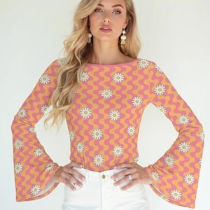 May include: A long-sleeved top featuring a pink and yellow wavy striped pattern with white daisy flower graphics. The garment has a boat neckline and flared sleeves, paired with plain white denim pants. The text Trendy Hip Buys is visible.