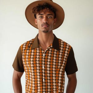 Pode incluir: Um chapéu fedora castanho e uma camisa abotoada de manga curta com um padrão pied-de-poule em tons de castanho, laranja e creme. A camisa tem gola e mangas castanhas escuras. O modelo está a usar calças de ganga azuis.