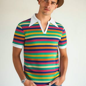 May include: A short-sleeved, striped polo shirt with a white collar and cuffs. The shirt features horizontal stripes in various colors, including red, yellow, green, and blue. The person is wearing a brown fedora hat and blue jeans.