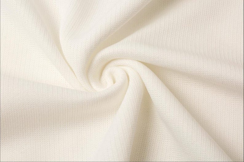 May include: Close-up of a cream-colored fabric with a textured weave. The fabric is draped and folded, creating soft shadows and highlights. The material appears to be lightweight and breathable, suitable for clothing or home decor.
