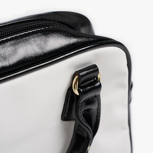 May include: A white and black handbag with a black leather handle and gold hardware. The bag has a zipper closure and a top handle.