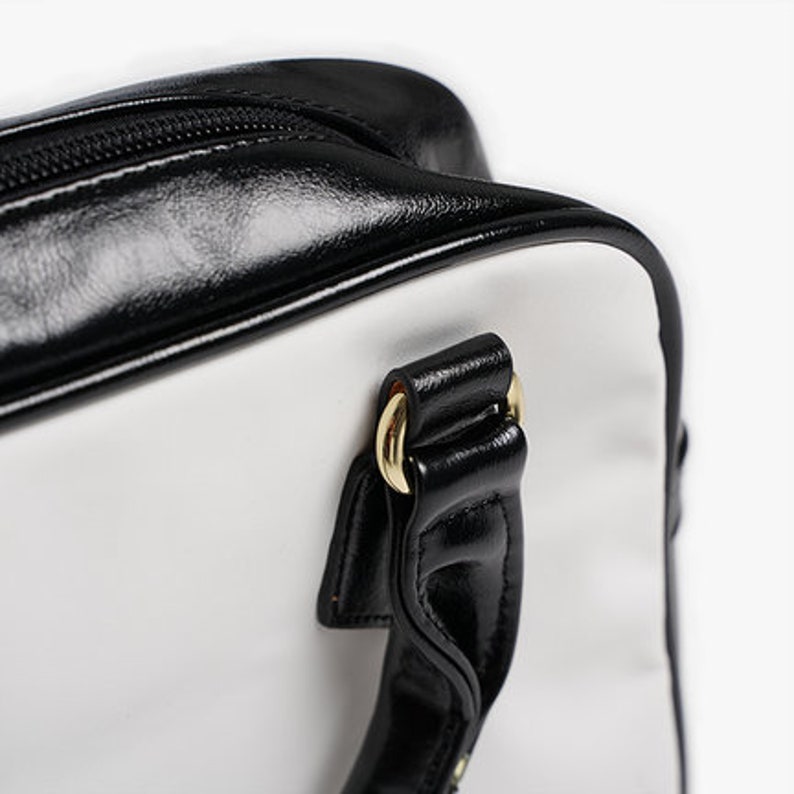May include: A white and black handbag with a black leather handle and gold hardware. The bag has a zipper closure and a top handle.