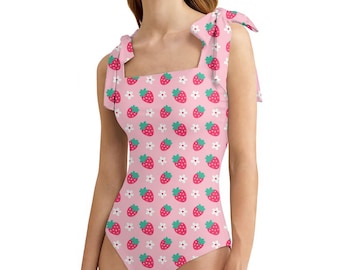 May include: A pink one-piece swimsuit with a strawberry and flower pattern. The swimsuit has tie straps and a square neckline. The strawberries are red with green leaves, and the flowers are white.