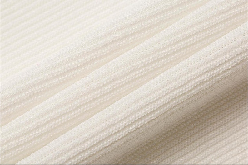 May include: Close-up of a textured, off-white fabric with a ribbed pattern. The fabric is draped, creating soft folds and shadows. The texture appears to be a knit or woven material, possibly used for clothing or home decor.