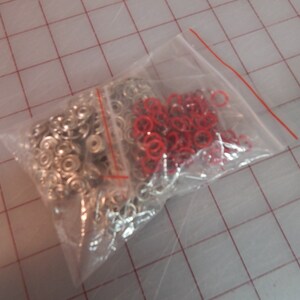 May include: A clear plastic bag filled with silver metal snap fasteners and red metal rings.
