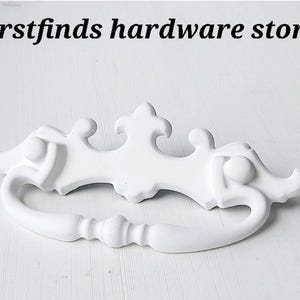 May include: A white, decorative drawer pull with a curved handle and ornate detailing. The hardware features a symmetrical design with a central flourish and is set against a white background. The text "firstfinds hardware store" is at the top.