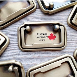 Peut inclure: Une collection de poignées de tiroir rectangulaires en métal couleur laiton avec un mécanisme de poignée tombante. La poignée centrale porte le texte firstfinds hardware store et une icône de feuille d'érable rouge.