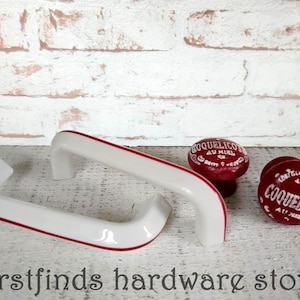 May include: White cabinet handles with a red trim and two red knobs with white text reading "Coquelicots au Miel". The hardware is displayed on a white surface in front of a white brick wall.