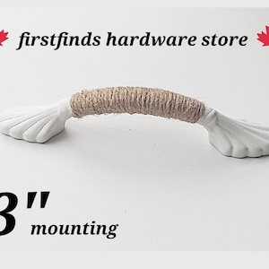 May include: A white cabinet pull with shell-shaped ends and a centre wrapped in natural tan jute twine. The text firstfinds hardware store and 3" mounting are visible. The handle features a curved design with a textured rope middle section.