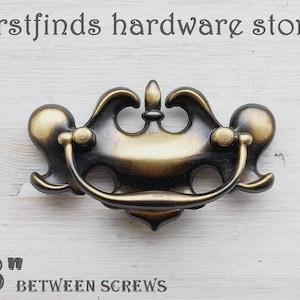 May include: An antique brass cabinet pull with a decorative scroll design. The pull has a D-shaped handle and is designed to be installed with two screws. The pull measures 3 inches between the screw holes.